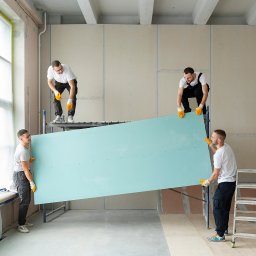 RENOVIO Sp. z o.o. - Czterech monterów w rękawicach unosi dużą, turkusową płytę gipsowo-kartonową na tle surowych ścian i rusztowań w jasnym, przestronnym wnętrzu.