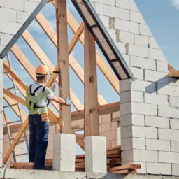 Budowa domu: murarz w kasku montuje drewnianą konstrukcję dachu na tle białych ścian z betonu komórkowego, widoczne niebo. Budowa w toku.