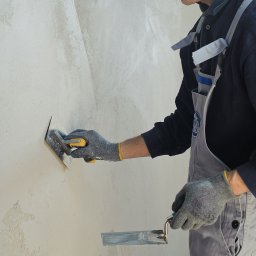 RENOVIO Sp. z o.o. - Tynkarz w szarym kombinezonie wyrównuje świeżą, jasną zaprawę na ścianie za pomocą kielni. Widoczne ślady po wcześniejszym nakładaniu tynku.