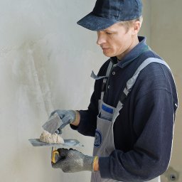 RENOVIO Sp. z o.o. - Tynkarz w kombinezonie nakłada zaprawę na ścianę za pomocą szpachelki i kielni. Widoczne nierówności na ścianie przed dalszą obróbką.