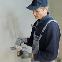 Tynkarz w kombinezonie nakłada zaprawę na ścianę za pomocą szpachelki i kielni. Widoczne nierówności na ścianie przed dalszą obróbką.