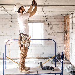 RENOVIO Sp. z o.o. - Elektryk w kasku, na rusztowaniu, montuje przewody elektryczne w surowym wnętrzu budynku z białych bloczków, z narzędziami przy pasie i wiertarką obok.