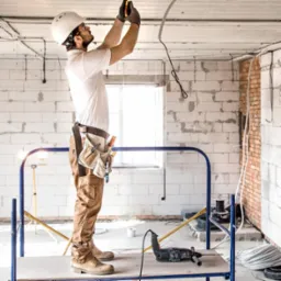 Elektryk w kasku, na rusztowaniu, montuje przewody elektryczne w surowym wnętrzu budynku z białych bloczków, z narzędziami przy pasie i wiertarką obok.