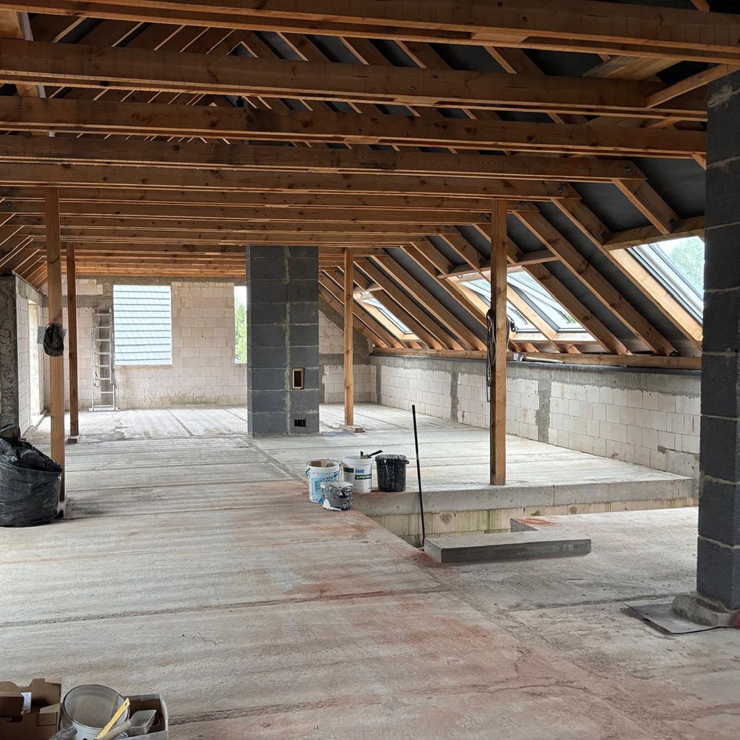 Poddasze w trakcie adaptacji: surowe ściany z betonu komórkowego, drewniana konstrukcja dachu z oknami połaciowymi, widoczne materiały budowlane i narzędzia.