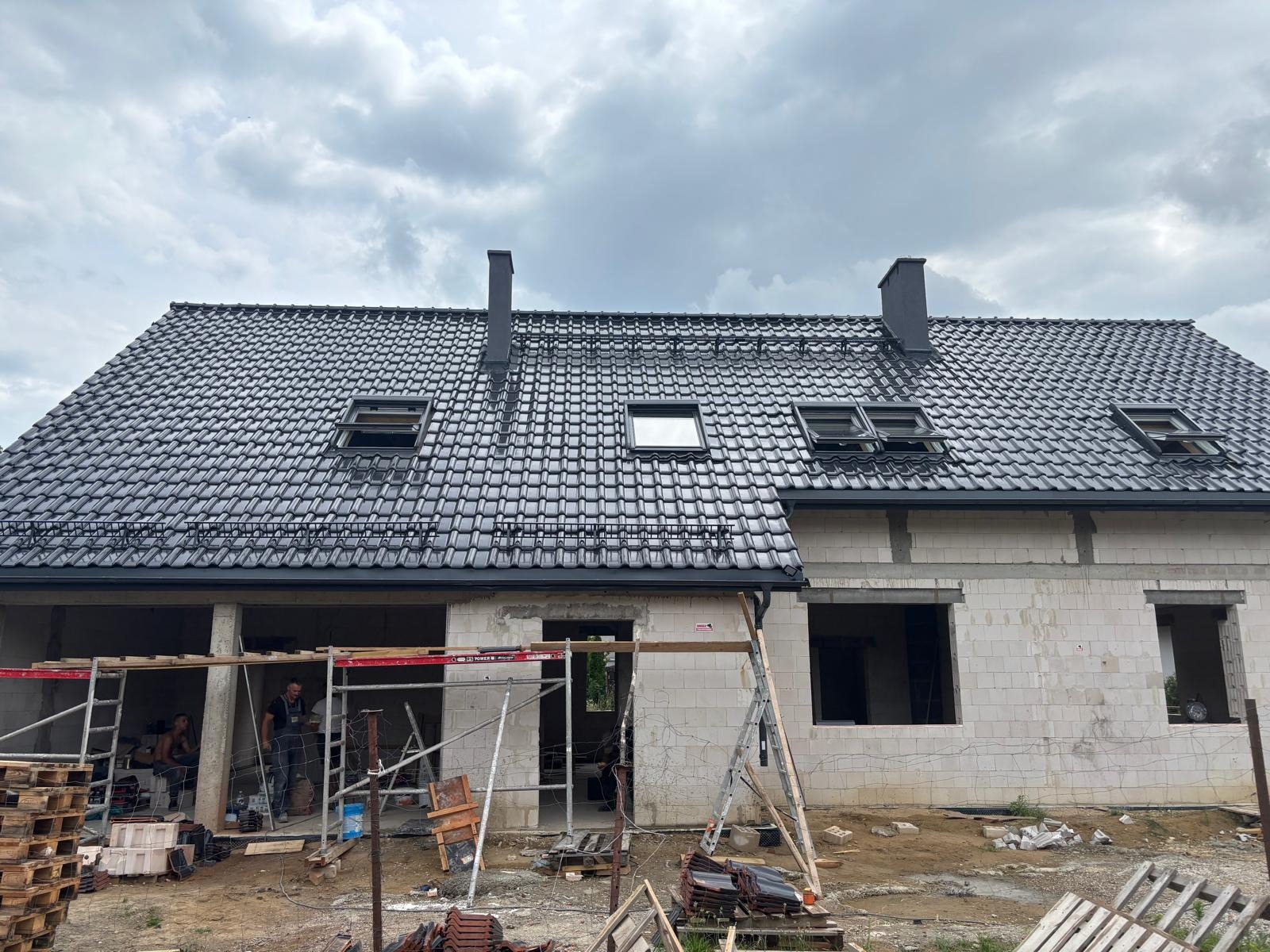 Dom w trakcie budowy z czarną dachówką, widoczne okna dachowe i rusztowania. Część budynku w stanie surowym, druga część z gotowym dachem. Pochmurne niebo w tle.