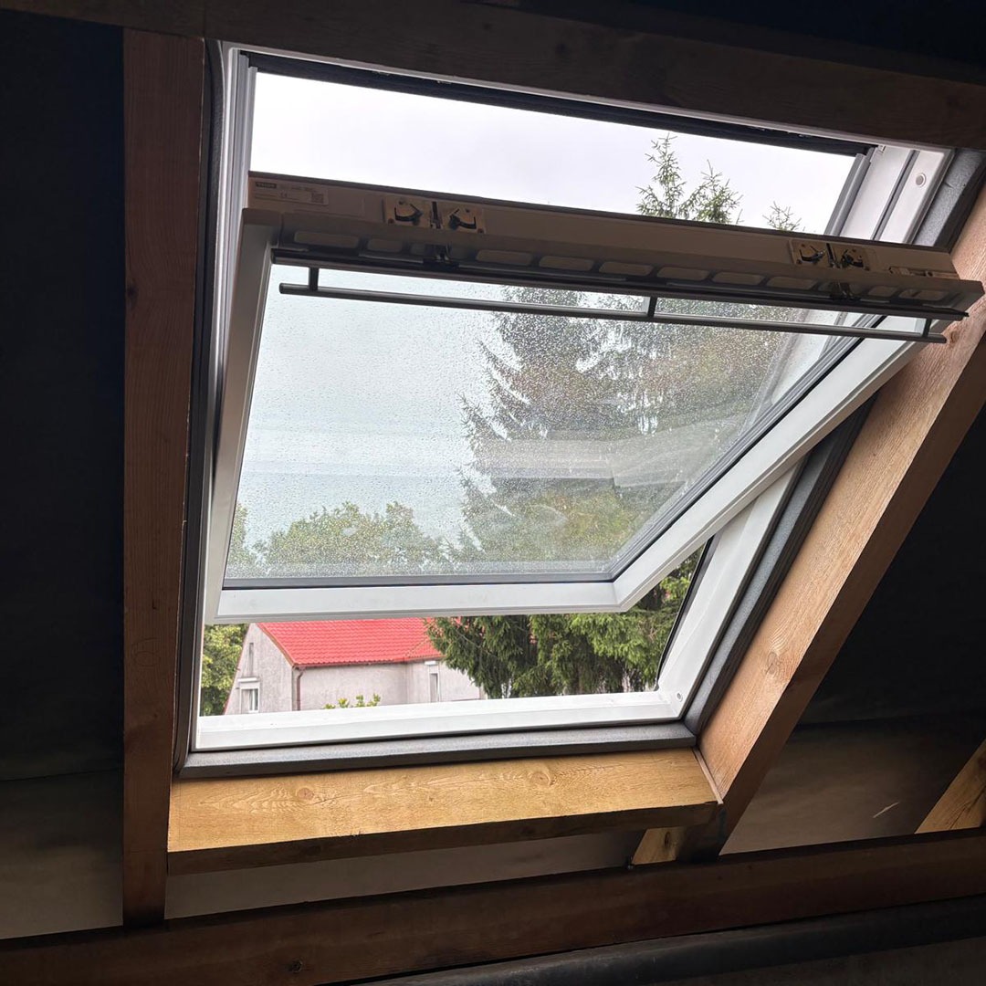 Otwarty, biały okno dachowe z widokiem na zewnątrz w deszczowy dzień. Widoczny czerwony dach domu i drzewa. Drewniane elementy konstrukcyjne poddasza.