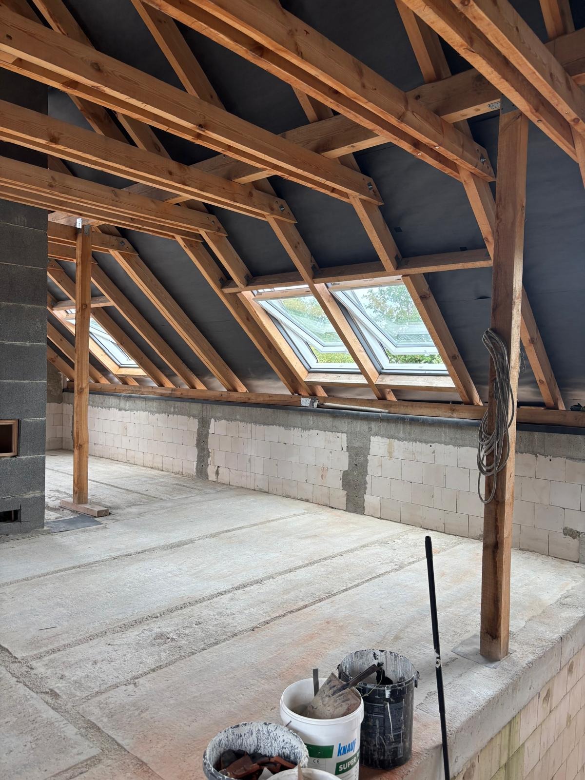 Poddasze w stanie surowym otwartym: drewniana konstrukcja dachu z oknami połaciowymi, ściany z białych bloczków, betonowa wylewka. Widoczne narzędzia i materiały budowlane.
