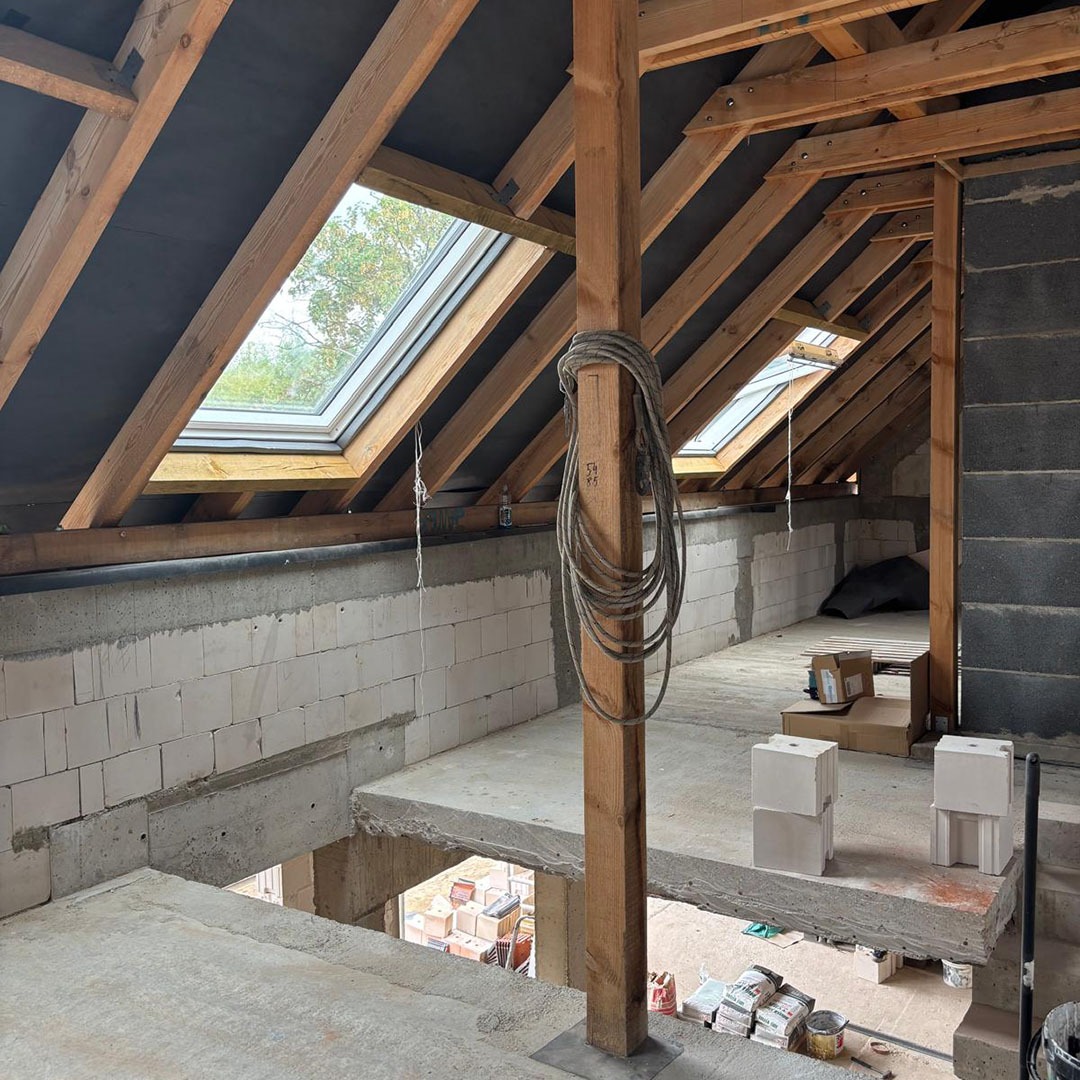 Poddasze w trakcie adaptacji: widoczne okna dachowe, drewniana konstrukcja więźby, murowane ściany z białych bloczków, betonowa wylewka. Na podłodze materiały budowlane.