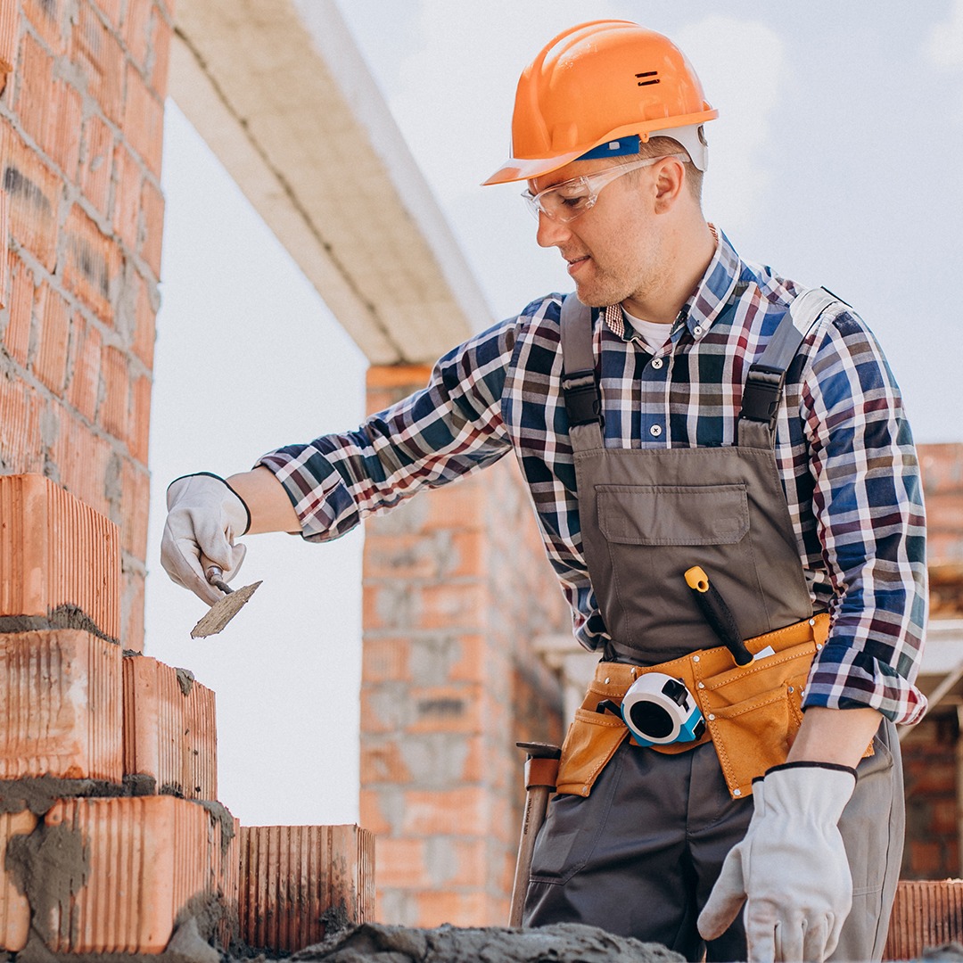 Murarz w kasku i okularach ochronnych, w trakcie murowania ściany z czerwonej cegły z użyciem kielni. Widoczne narzędzia w pasie.