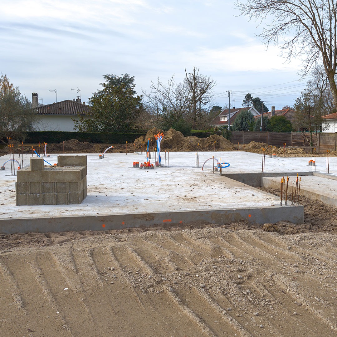 Wylana betonowa płyta fundamentowa z wystającymi prętami zbrojeniowymi i instalacjami, przygotowana pod budowę domu jednorodzinnego. Widok na plac budowy w pochmurny dzień.