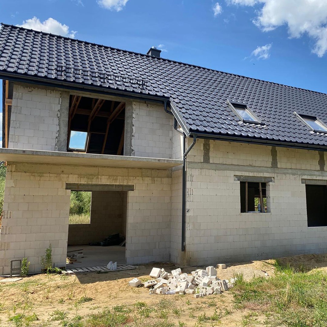 Dom w stanie surowym otwartym z czarną dachówką i oknami dachowymi. Widoczne białe bloczki i niezadaszony otwór wejściowy. Częściowo ułożone bloczki na ziemi.
