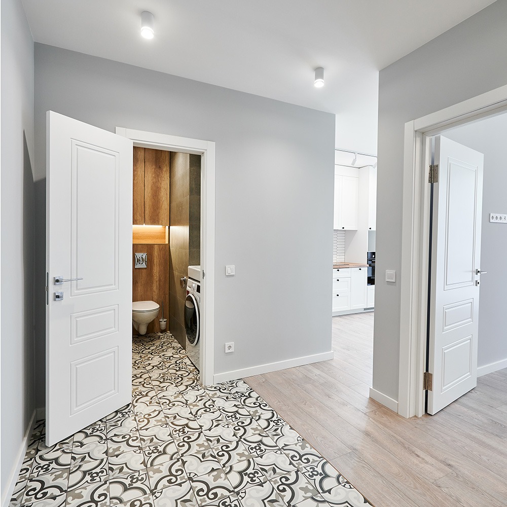 Wnętrze mieszkania: wzorzysta podłoga w korytarzu, otwarte drzwi do łazienki z drewnianymi akcentami i pralką, w tle widoczna biała kuchnia.