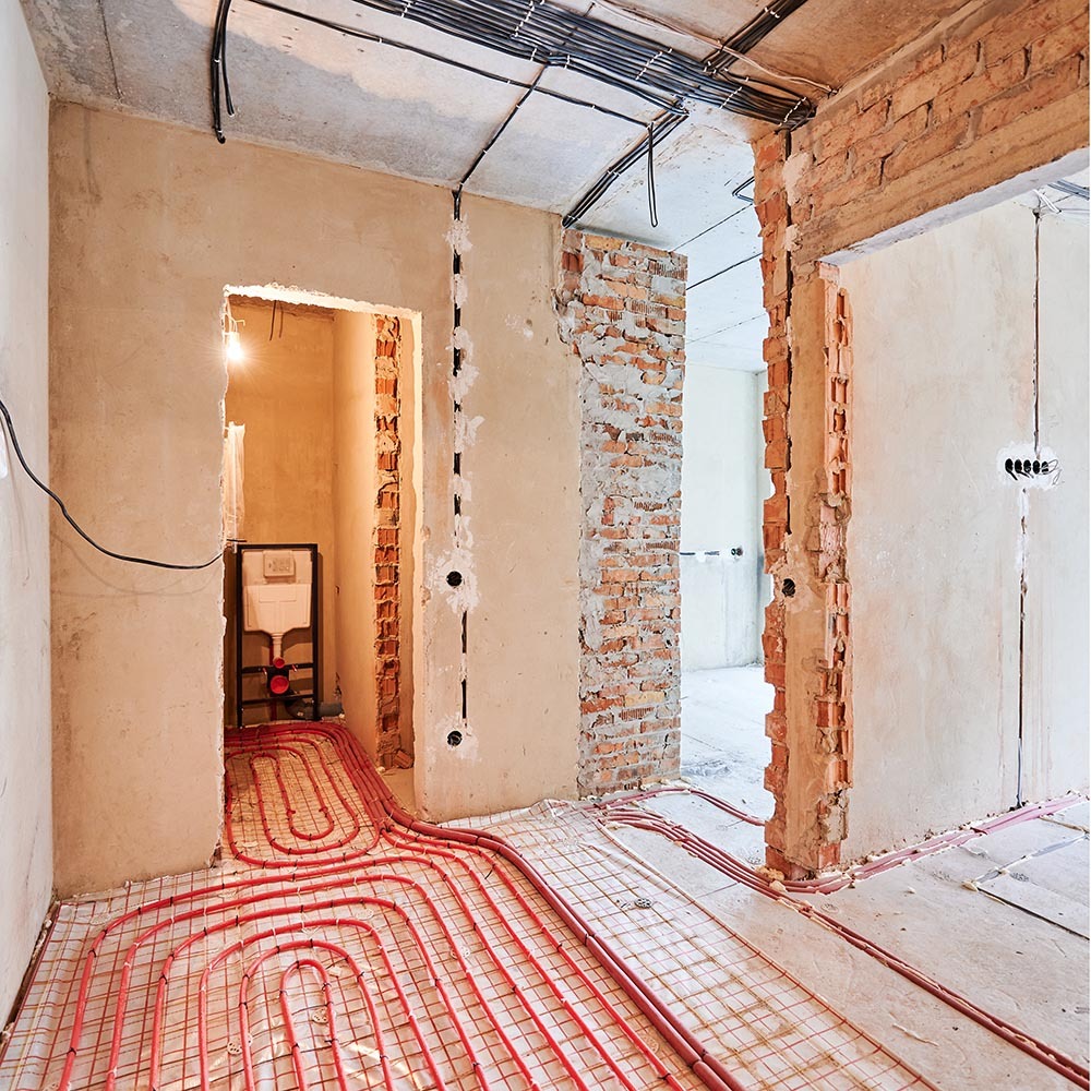Instalacja ogrzewania podłogowego z rurami w kolorze czerwonym, widok na surowy stan ścian z cegły i betonu, przygotowanie do dalszych prac wykończeniowych.