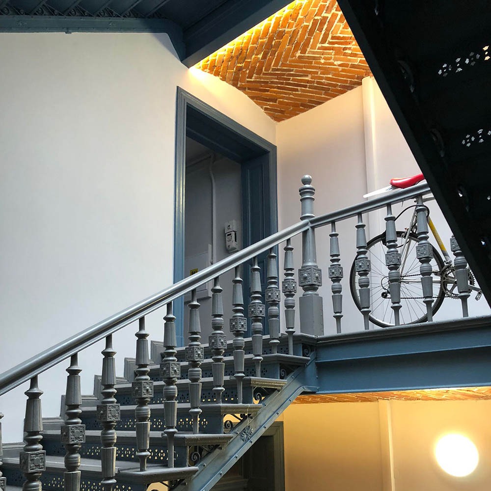 Klatka schodowa z szarymi, zdobionymi schodami i balustradą. W tle mur z cegły na suficie i rower oparty o ścianę. Drzwi z niebieską framugą.