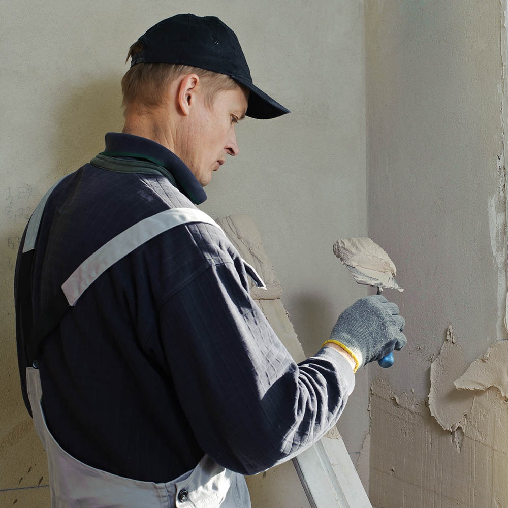 Mężczyzna w czapce, z kielnią, tynkuje ścianę w rogu pokoju. Świeży tynk w jasnym odcieniu. Fragment drabiny oparty o ścianę.