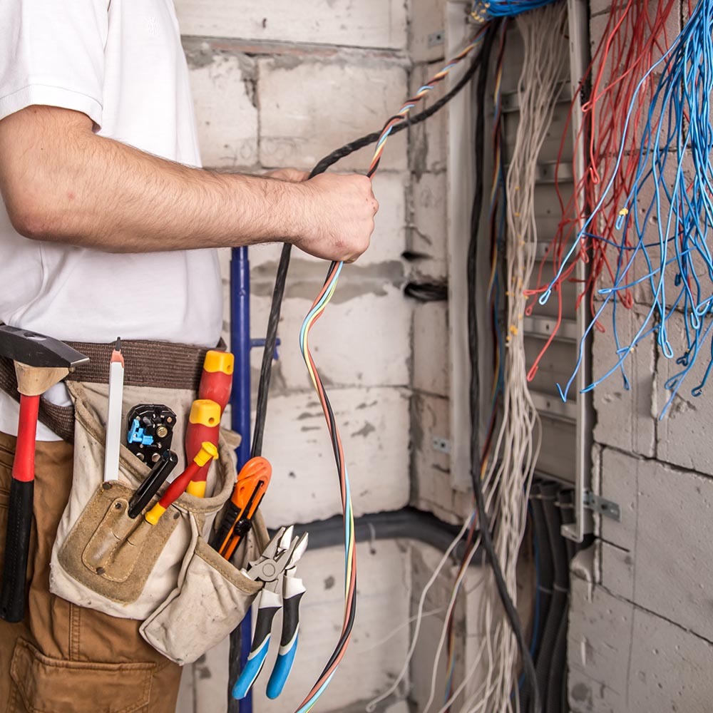 Elektryk w trakcie pracy: instalacja przewodów w surowym wnętrzu. Widoczne narzędzia w pasie i kolorowe kable. Ściany z białych pustaków.