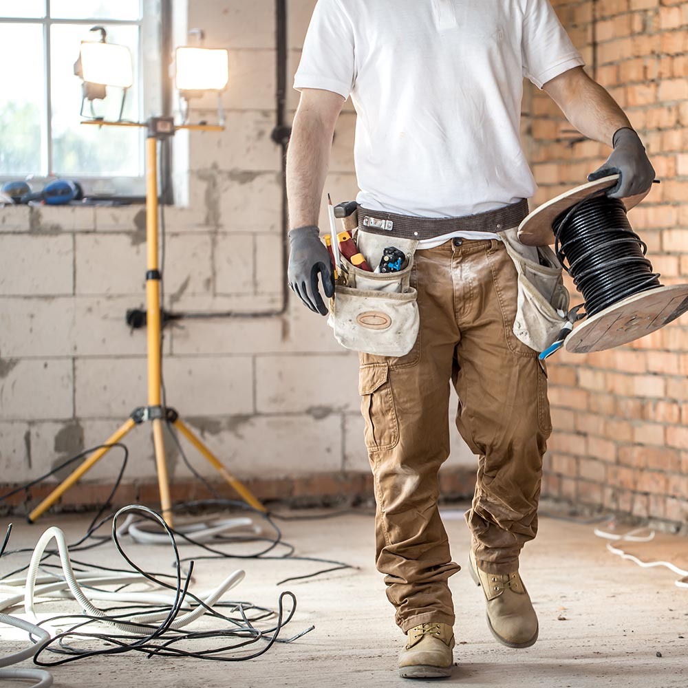 Elektryk z narzędziami i kablem na szpuli, w surowym wnętrzu z ceglaną ścianą i lampą budowlaną. Widoczne kable na podłodze, w trakcie prac instalacyjnych.