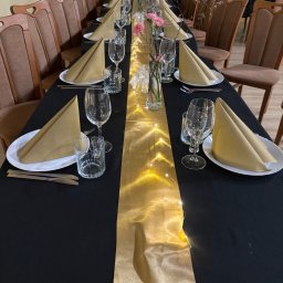 AG MIL Dominika - Uroczysty stół z czarnym obrusem, złotym bieżnikiem z lampkami, nakryciami i kwiatami. Aranżacja na przyjęcie z brązowymi krzesłami w tle.