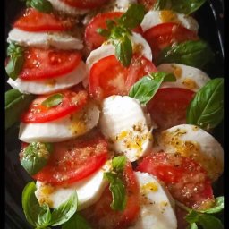 AG MIL Dominika - Caprese na czarnym talerzu: plastry pomidorów i mozzarelli przełożone bazylią, skropione oliwą. Widok z góry, naturalne światło.