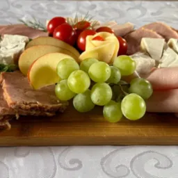 Dekoracyjna deska z wędlinami, serem brie, jabłkami, winogronami i pomidorami koktajlowymi na białym obrusie. Ser uformowany w kształt róży.