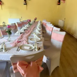 Uroczysty stół z dekoracjami w odcieniach różu: kwiaty, serwetki, wstążki na krzesłach. Przygotowania do przyjęcia w przestronnym wnętrzu z żółtymi ścianami.