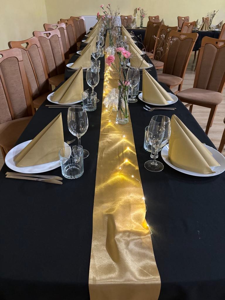 Uroczysty stół z czarnym obrusem, złotym bieżnikiem z lampkami, nakryciami i kwiatami. Aranżacja na przyjęcie z brązowymi krzesłami w tle.