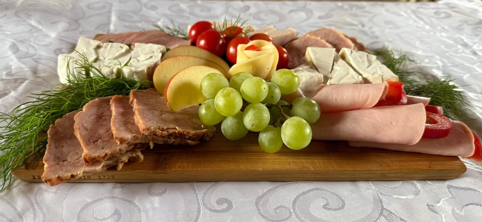 Dekoracyjna deska z wędlinami, serem brie, jabłkami, winogronami i pomidorami koktajlowymi na białym obrusie. Ser uformowany w kształt róży.