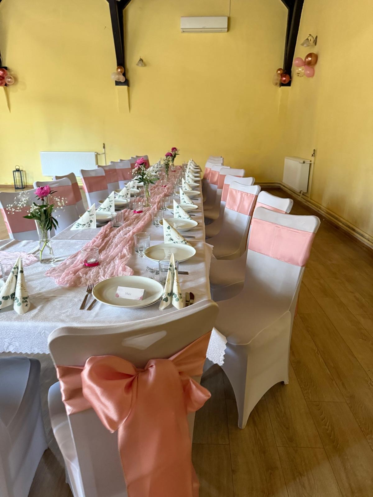 Uroczysty stół z dekoracjami w odcieniach różu: kwiaty, serwetki, wstążki na krzesłach. Przygotowania do przyjęcia w przestronnym wnętrzu z żółtymi ścianami.