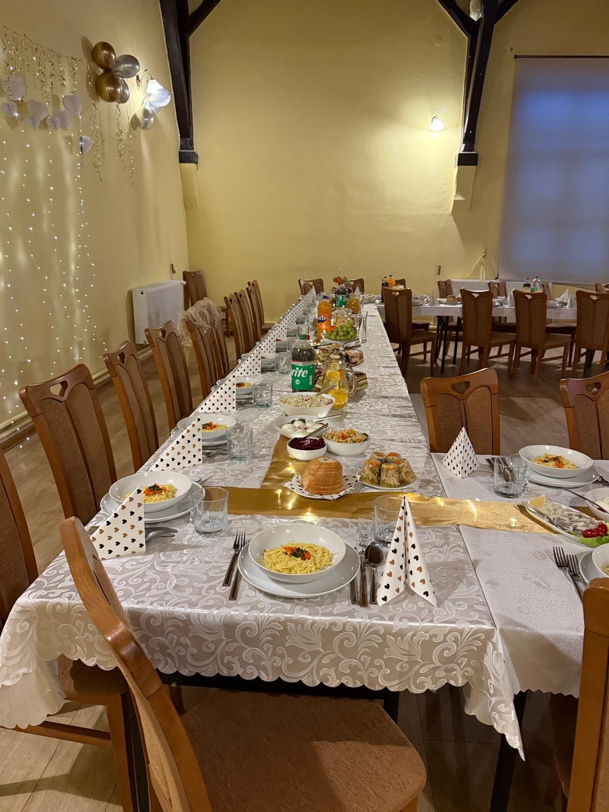 Długi stół z nakryciami i jedzeniem, dekorowany złotym bieżnikiem i balonami, gotowy na przyjęcie gości. Widoczne napoje, sałatki i dania główne.