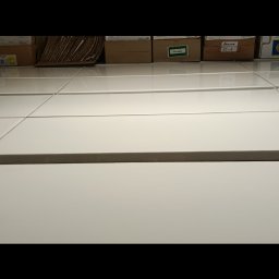 sandra pustelnik - Białe, błyszczące płytki podłogowe lub ścienne, ułożone w regularną siatkę, z kartonami w tle. Widoczne refleksy światła na powierzchni płytek.