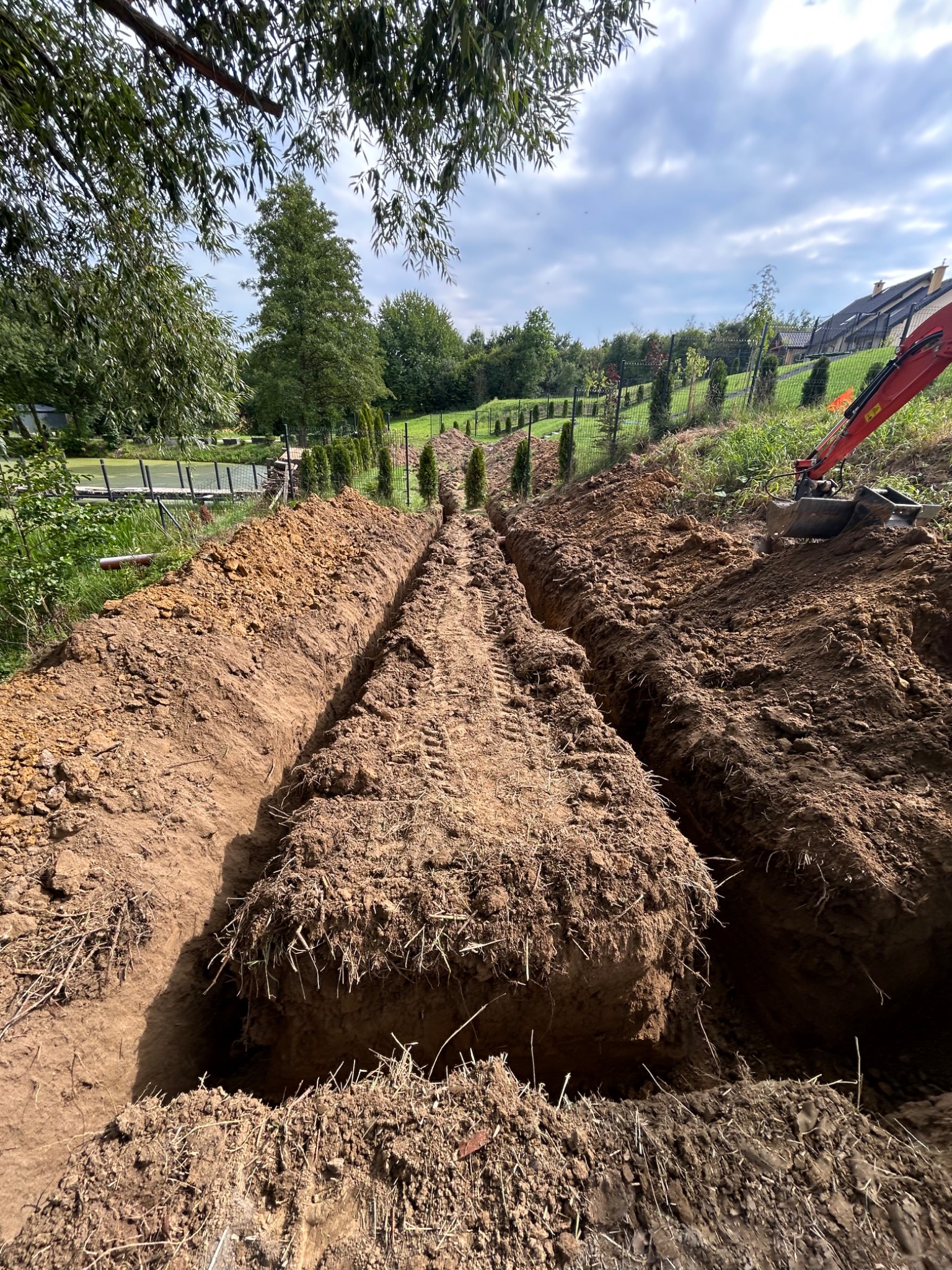 Długie, równoległe wykopy w ziemi, prawdopodobnie pod instalacje, wykonane na terenie zielonym z widocznym fragmentem koparki i drzewami w tle.