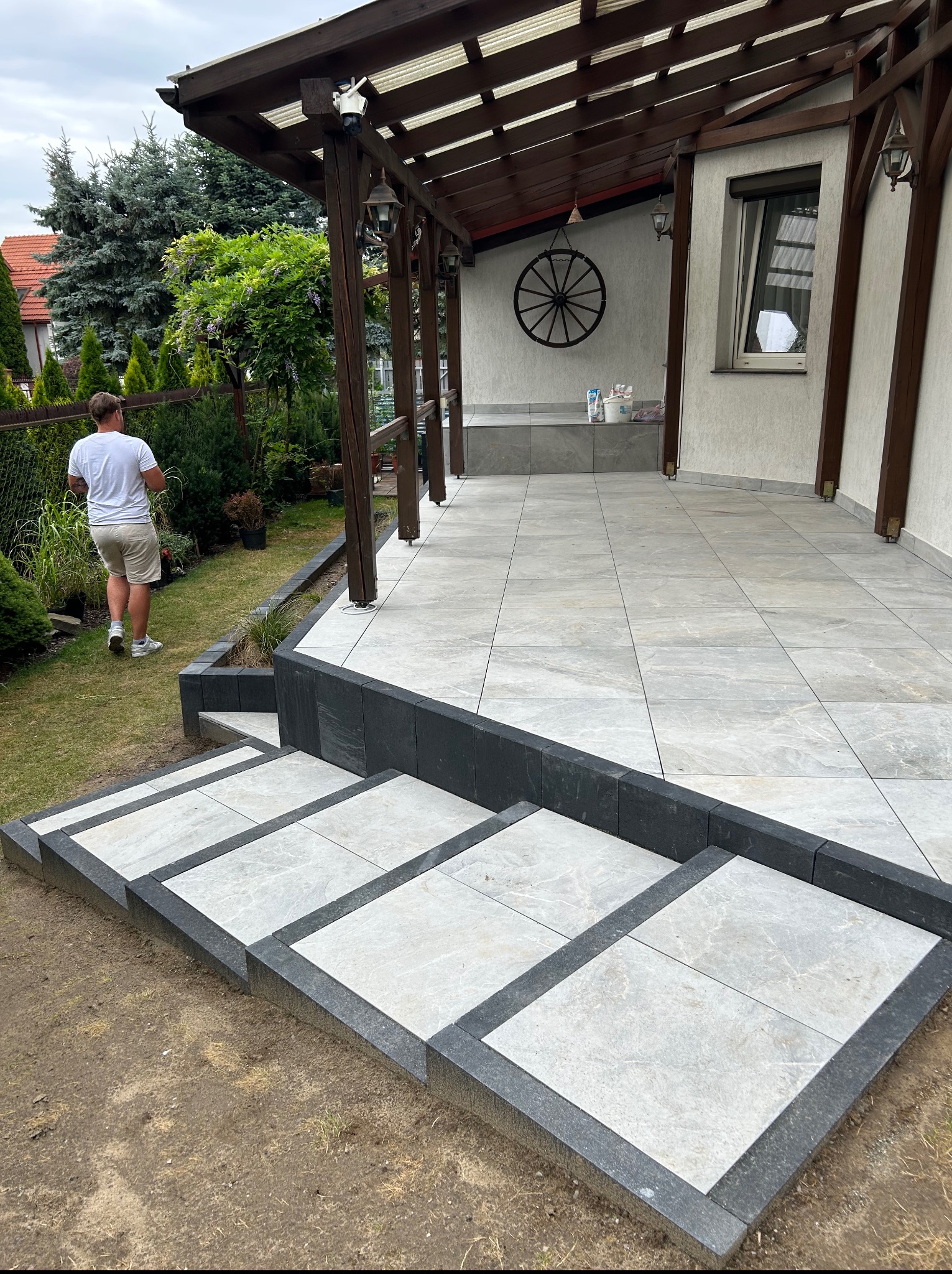 Nowoczesny taras z jasnoszarych płyt z ciemnoszarym obramowaniem schodów, zadaszenie z drewnianą konstrukcją, ogród w tle i osoba w oddali.