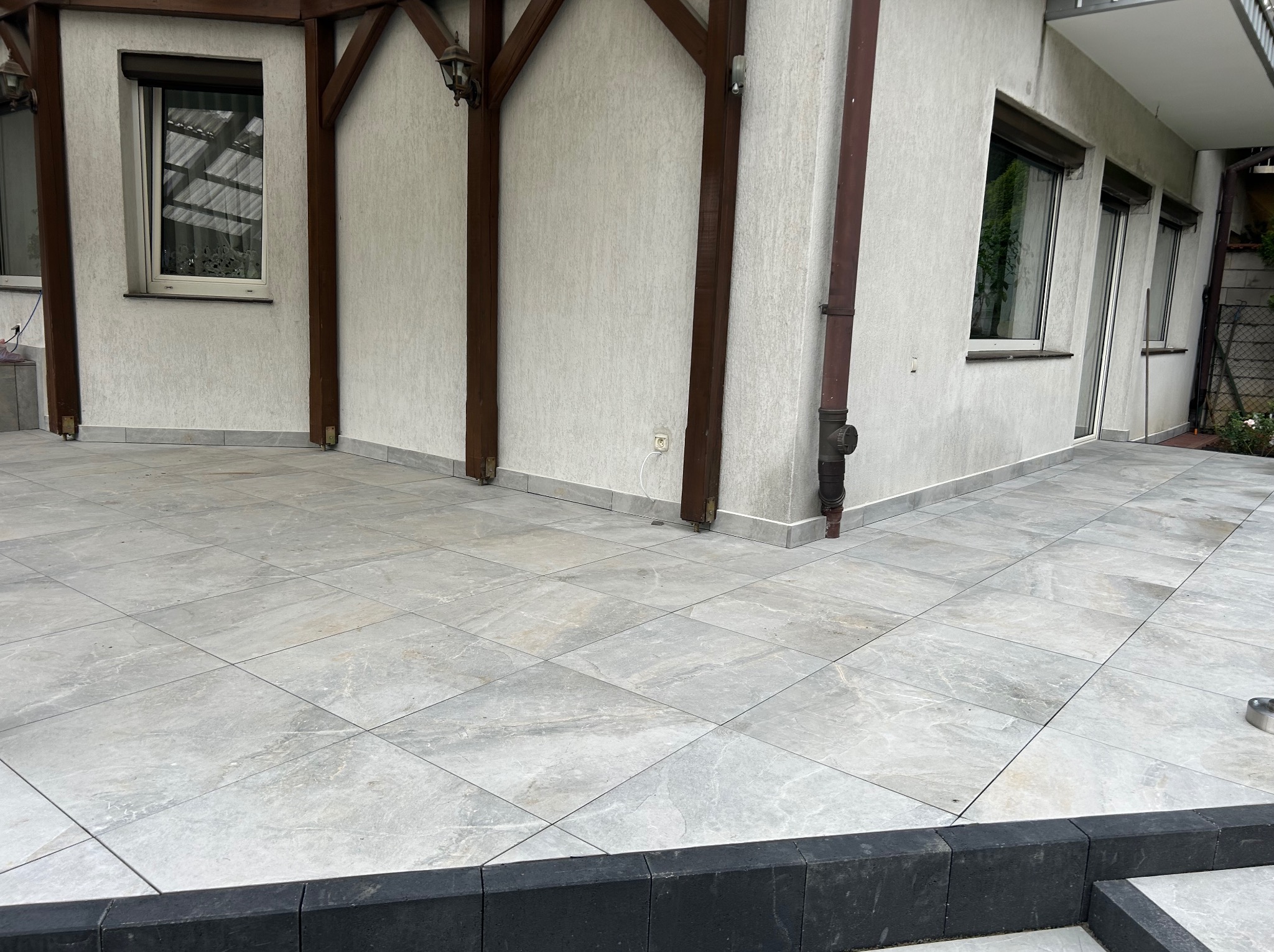 Jasnoszara, elegancka posadzka tarasowa z płytek ceramicznych ułożonych w karo, z widocznym fragmentem elewacji budynku i drewnianej konstrukcji zadaszenia.