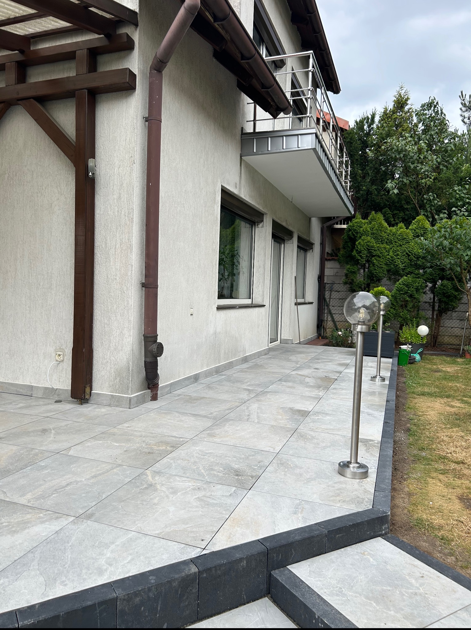 Jasnoszara, nowa nawierzchnia z płytek na zewnątrz domu, z widocznymi lampami ogrodowymi i fragmentem balkonu z metalową barierką. Ujęcie z niskiego kąta.