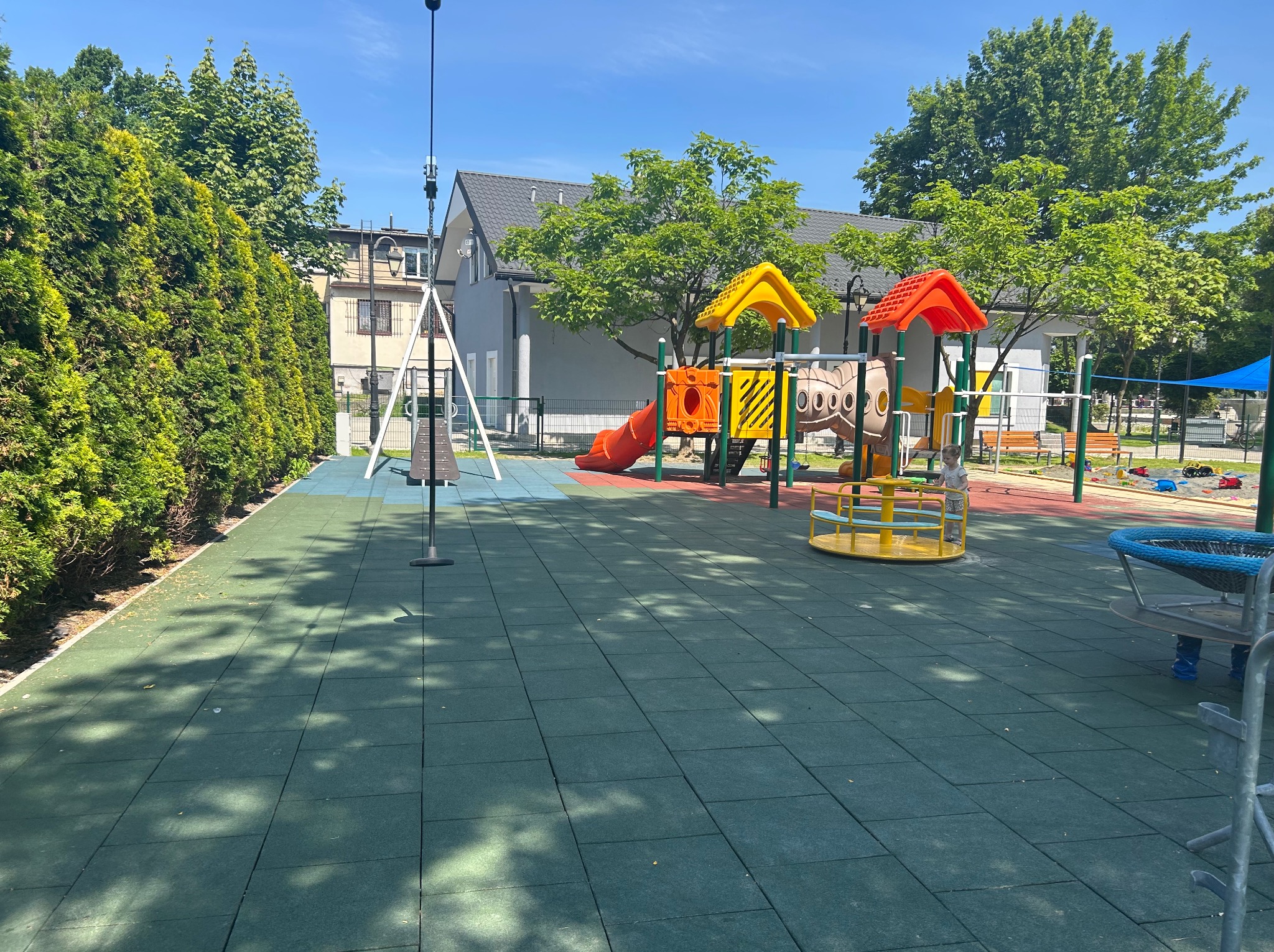 Kolorowy plac zabaw z bezpieczną, gumową nawierzchnią i różnorodnymi urządzeniami, otoczony zielenią, w słoneczny dzień. Widoczne elementy małej architektury.