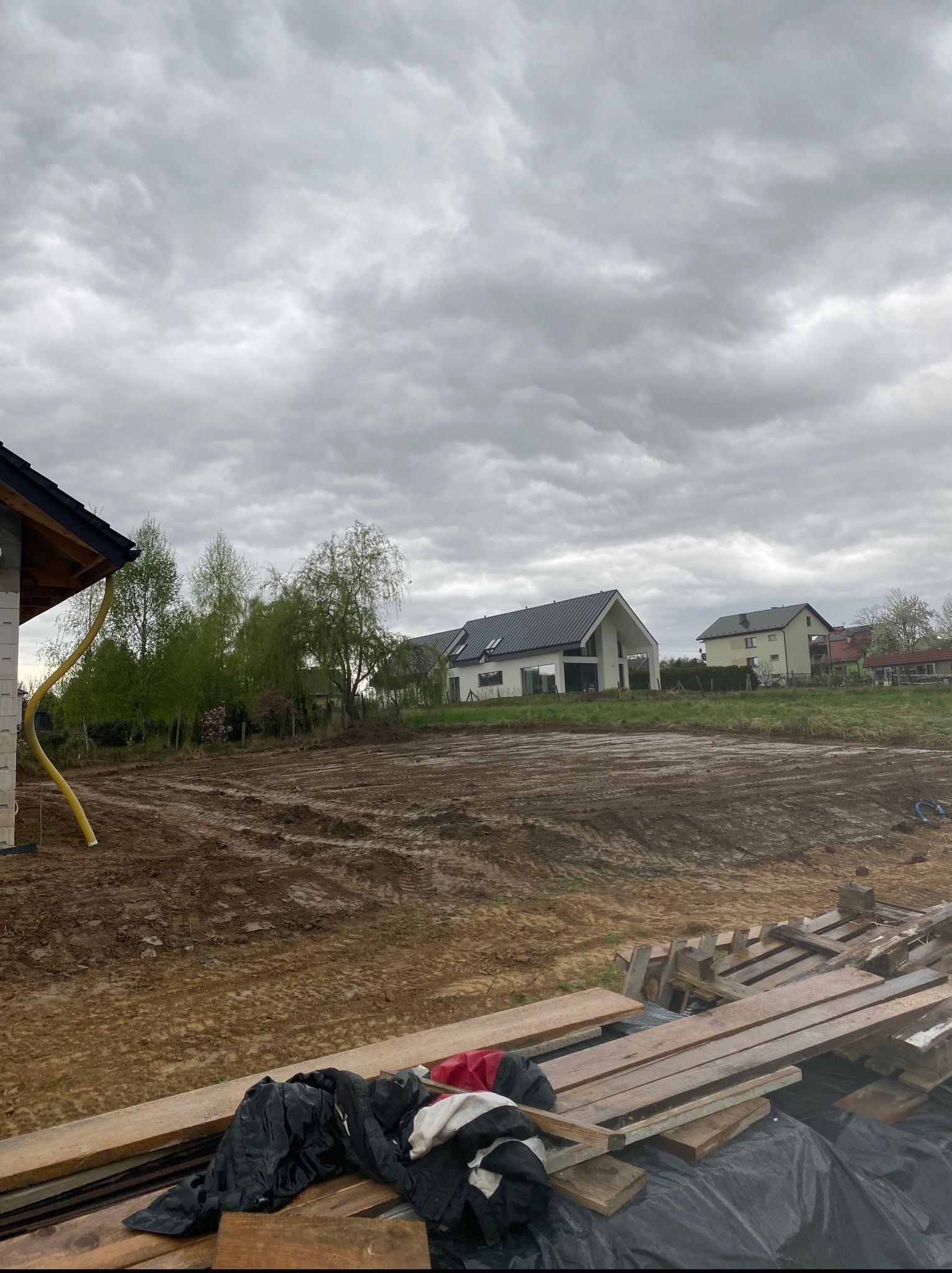 Przygotowany, wyrównany teren pod budowę domu jednorodzinnego, widoczne ślady opon maszyn budowlanych, w tle zabudowa jednorodzinna i pochmurne niebo.