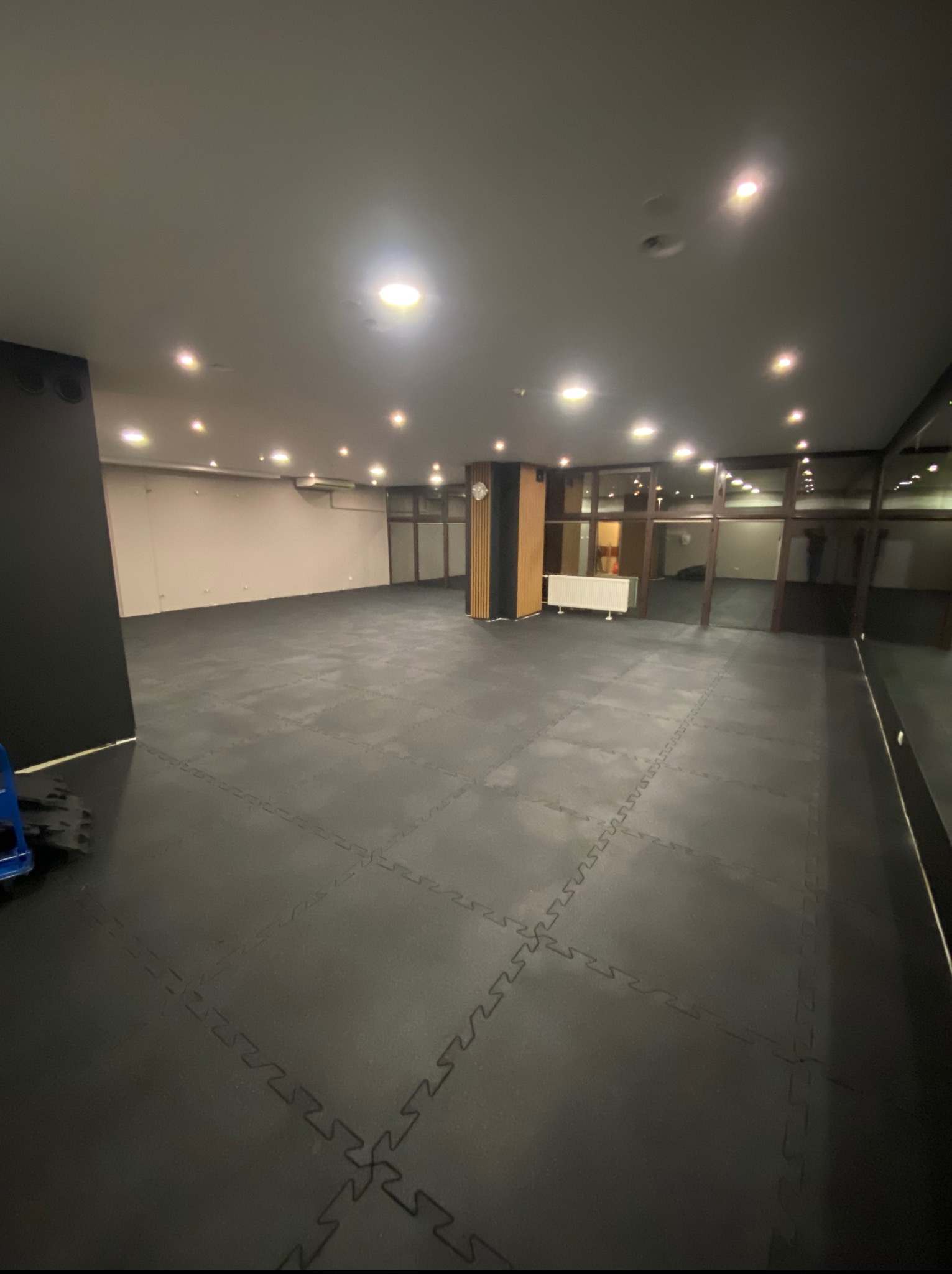 Przestronne wnętrze z czarną podłogą z puzzli, jasnymi ścianami i oknami. Widoczne oświetlenie sufitowe i fragment grzejnika. Ujęcie z niskiego kąta podkreśla głębię pomieszczenia.