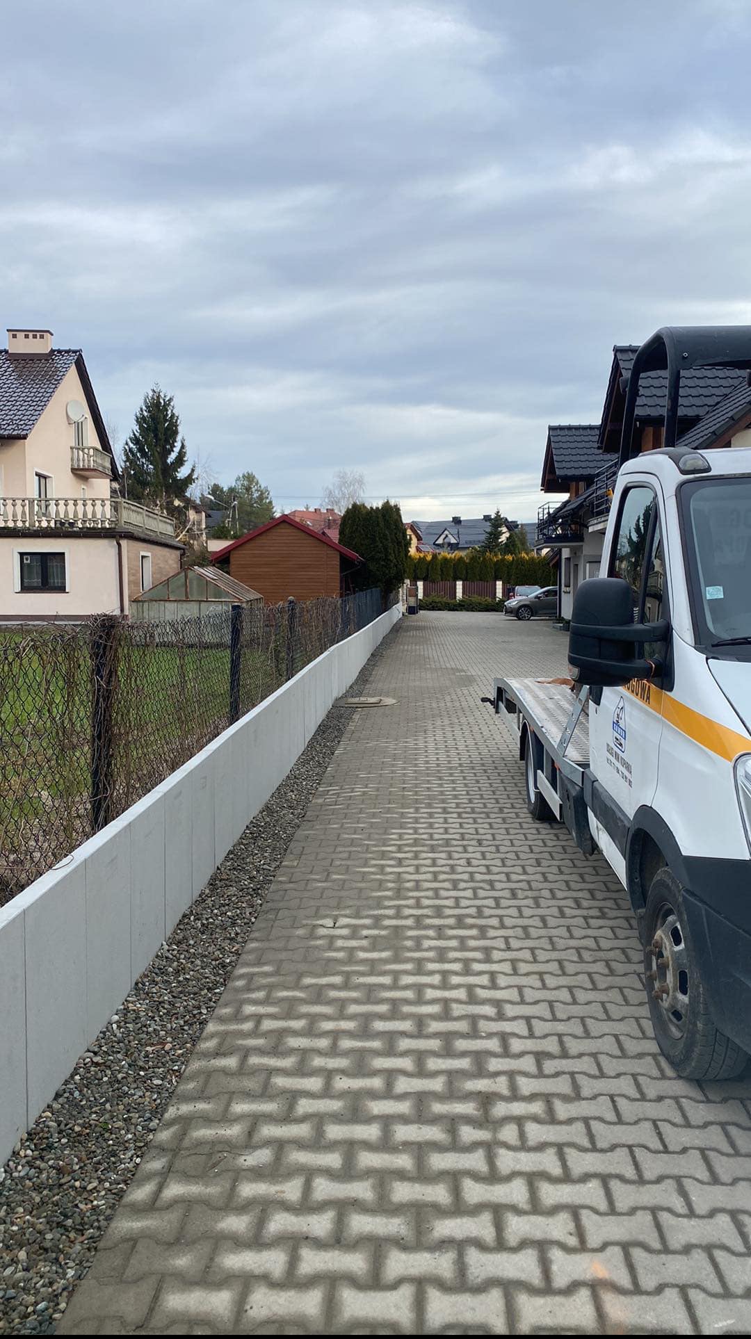 Nowy, betonowy murek oporowy oddziela posesję od drogi z kostki. Na drugim planie domy i auto dostawcze z platformą. Pochmurne niebo.