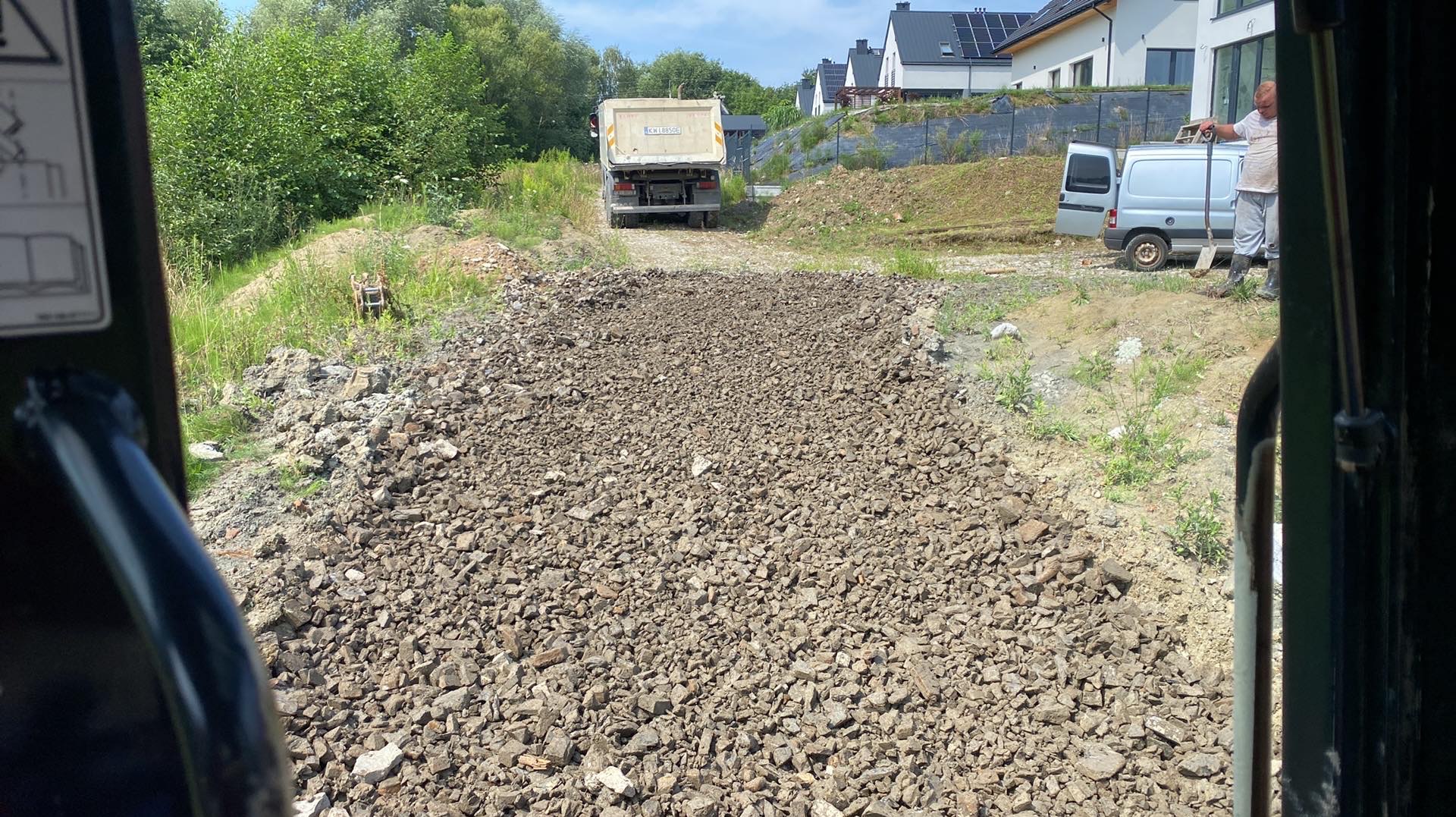 Utwardzony teren gruzem, widok z kabiny maszyny. W tle ciężarówka i budynki mieszkalne. Pracownik z łopatą obok samochodu dostawczego.