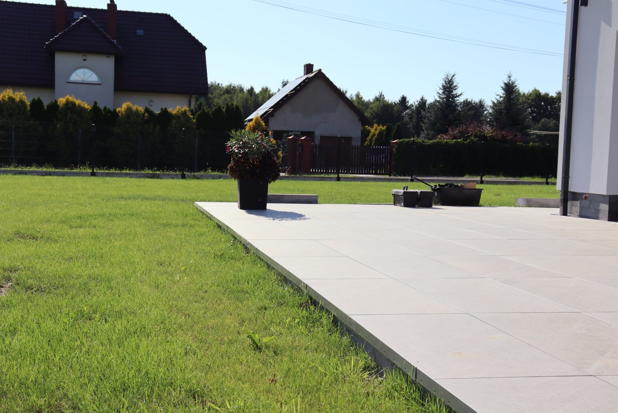 Nowoczesny taras z jasnych płyt betonowych, z donicą i narzędziami. W tle domy i zieleń, słoneczny dzień.