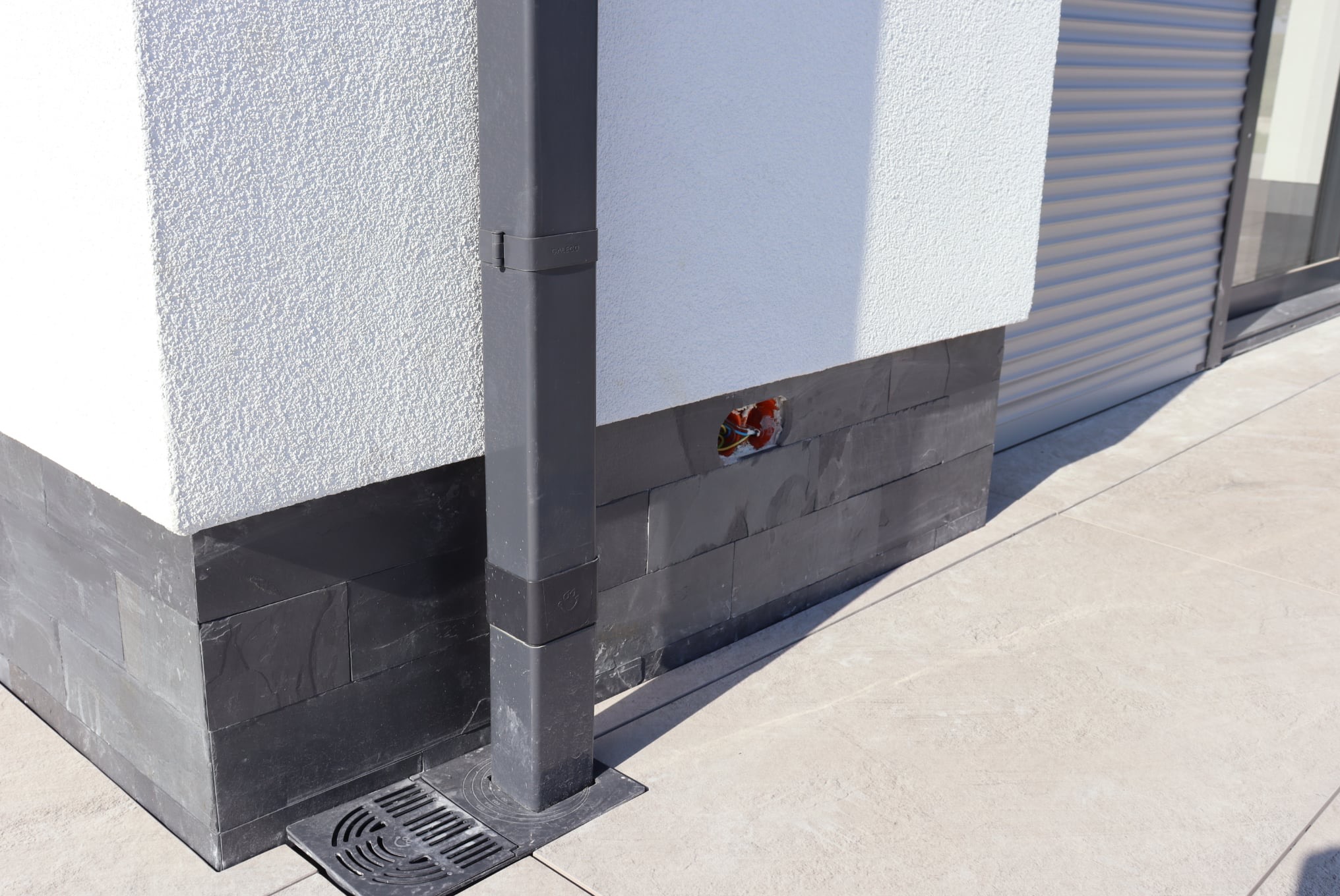 Szara rura spustowa z kratką drenażową przy elewacji z okładziną imitującą kamień. Widoczne przewody w puszce elektrycznej. Jasna kostka brukowa.