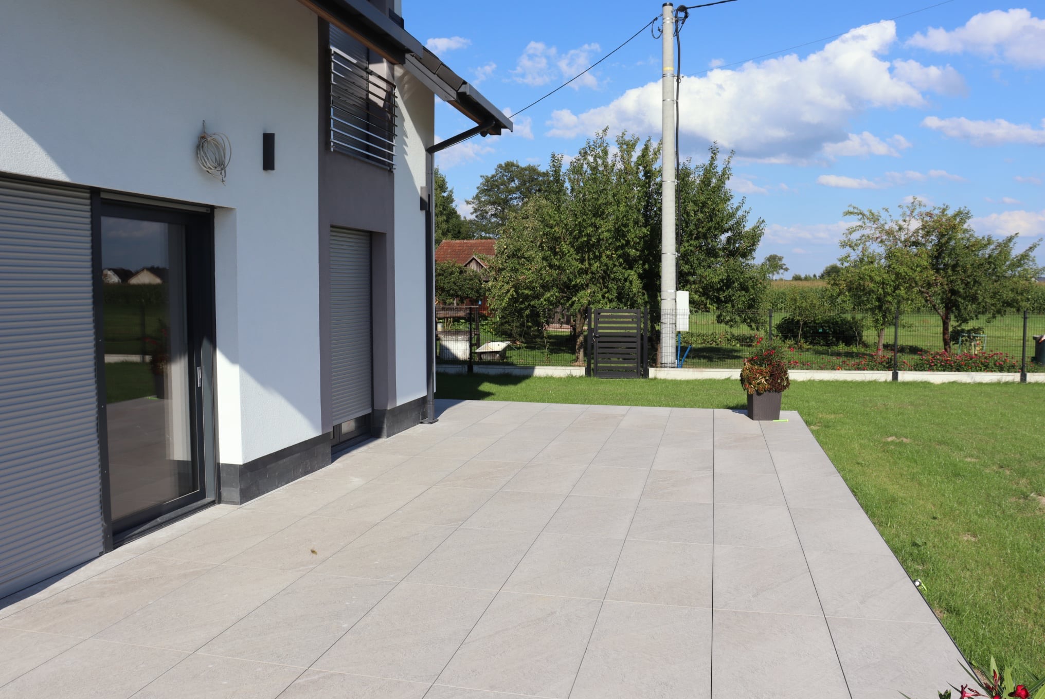 Jasnoszary taras z płytek przy nowoczesnym domu z szarymi roletami, widok na ogród z drzewami i słupem energetycznym w tle, słoneczny dzień.