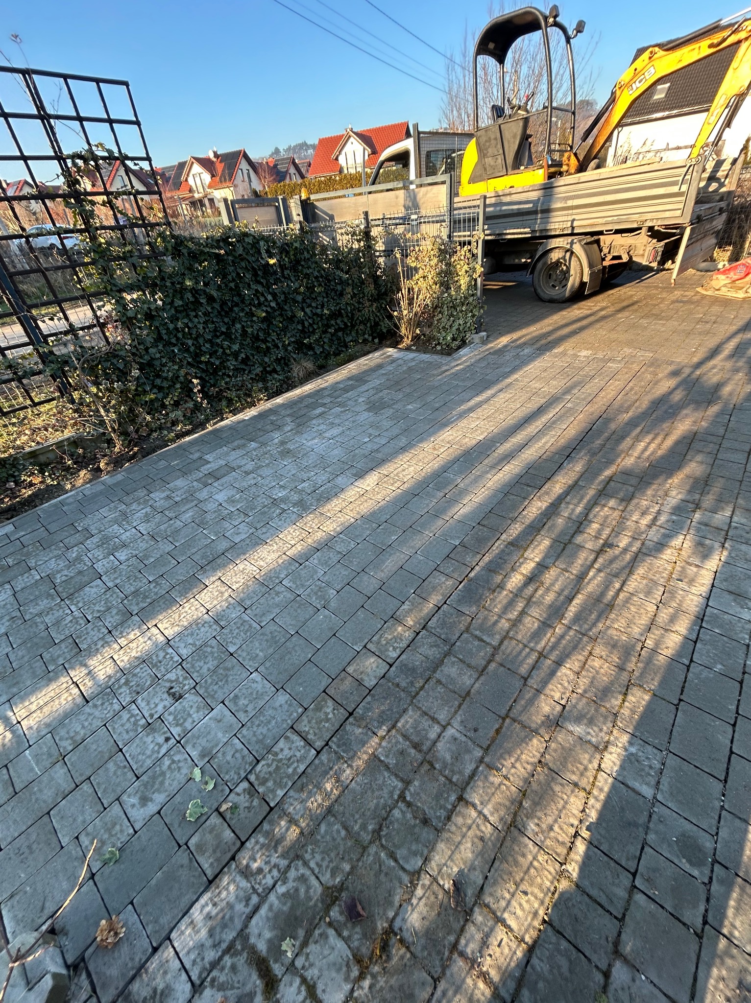 Ułożona kostka brukowa na podjeździe, z widocznym mini-koparką na przyczepie w tle. Długie cienie drzew padają na powierzchnię. W tle domy z czerwonymi dachami.