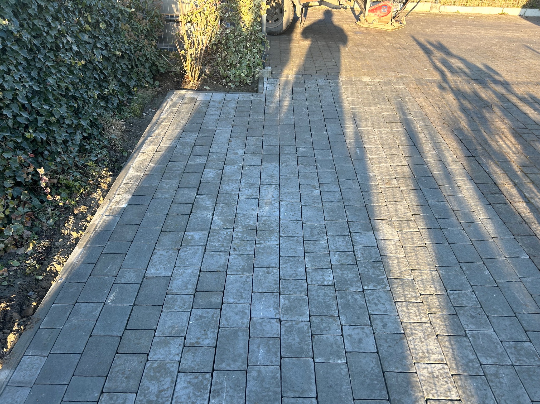 Ułożona szara kostka brukowa na podjeździe, cień osoby i maszyny w tle, obok żywopłot. Widok z góry na nową nawierzchnię.