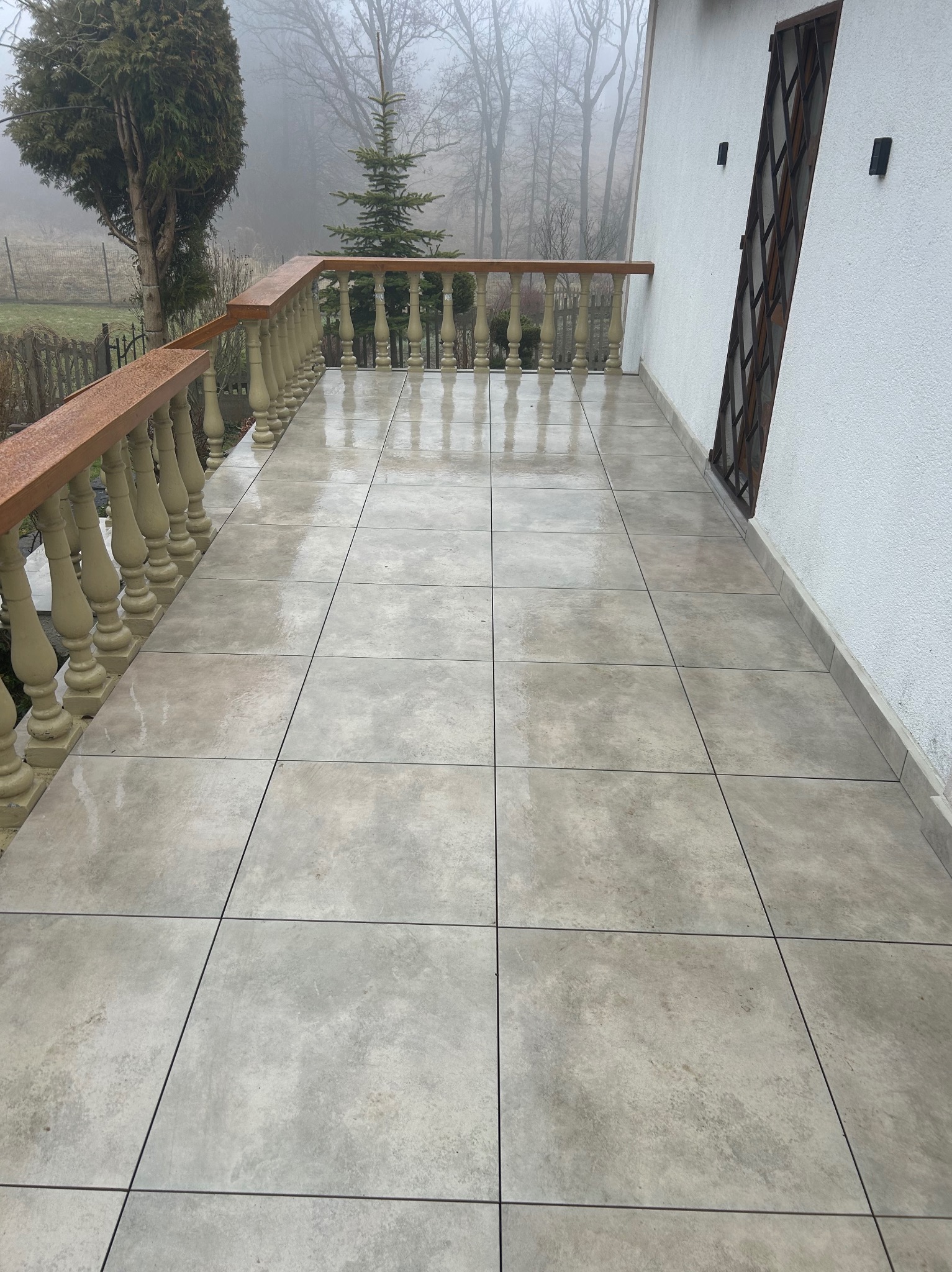 Balkon wyłożony jasnymi, kwadratowymi płytkami ceramicznymi z ciemnymi fugami. Balustrada z drewnianym poręczą i jasnymi, ozdobnymi słupkami. W tle drzewa we mgle.