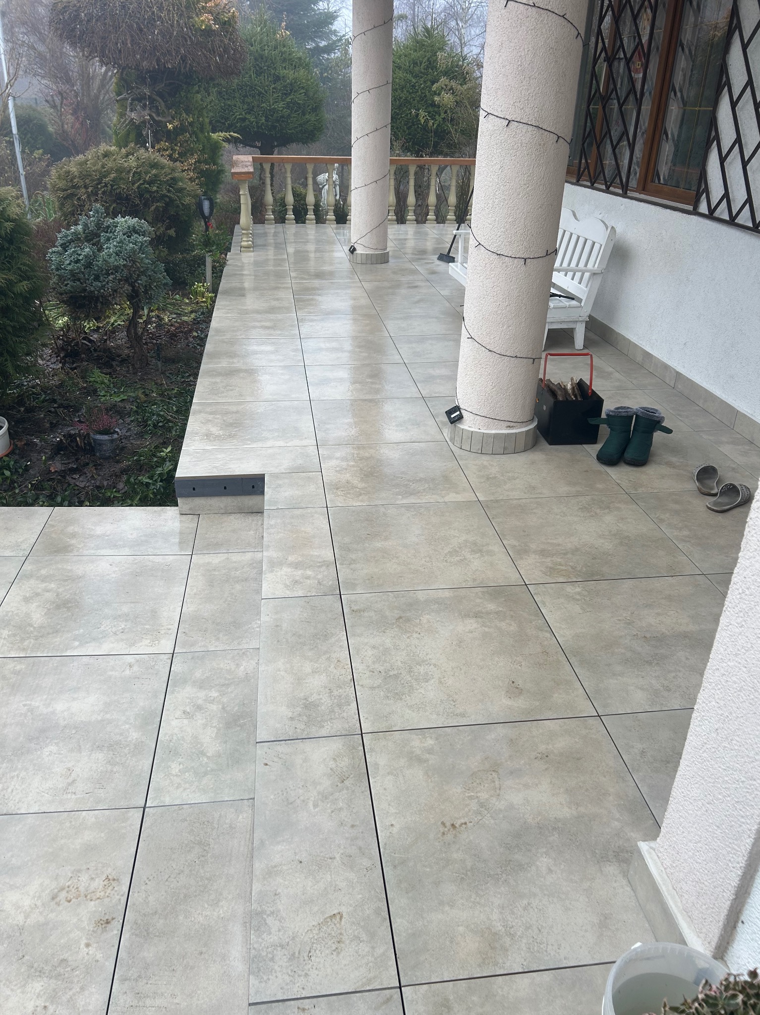 Jasne, kwadratowe płytki na zewnątrz budynku, tworzące taras z kolumnami. Widoczne buty, rośliny i elementy dekoracyjne. Ujęcie z góry, podkreślające wzór i fakturę płytek.