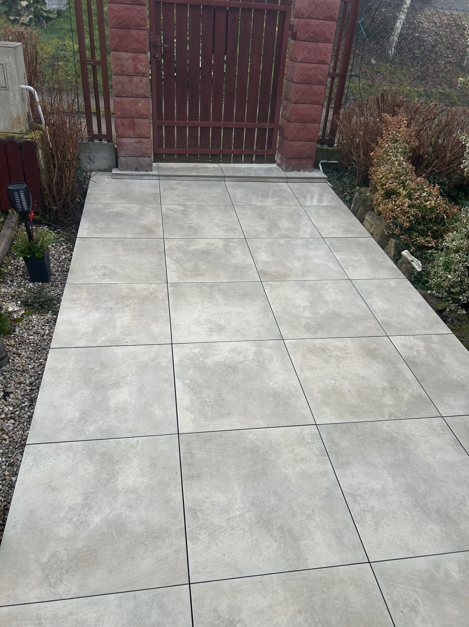 Ułożona ścieżka z jasnoszarych, kwadratowych płyt prowadząca do drewnianej bramki z ceglanymi słupkami. Po bokach widoczne rośliny i kamyki.