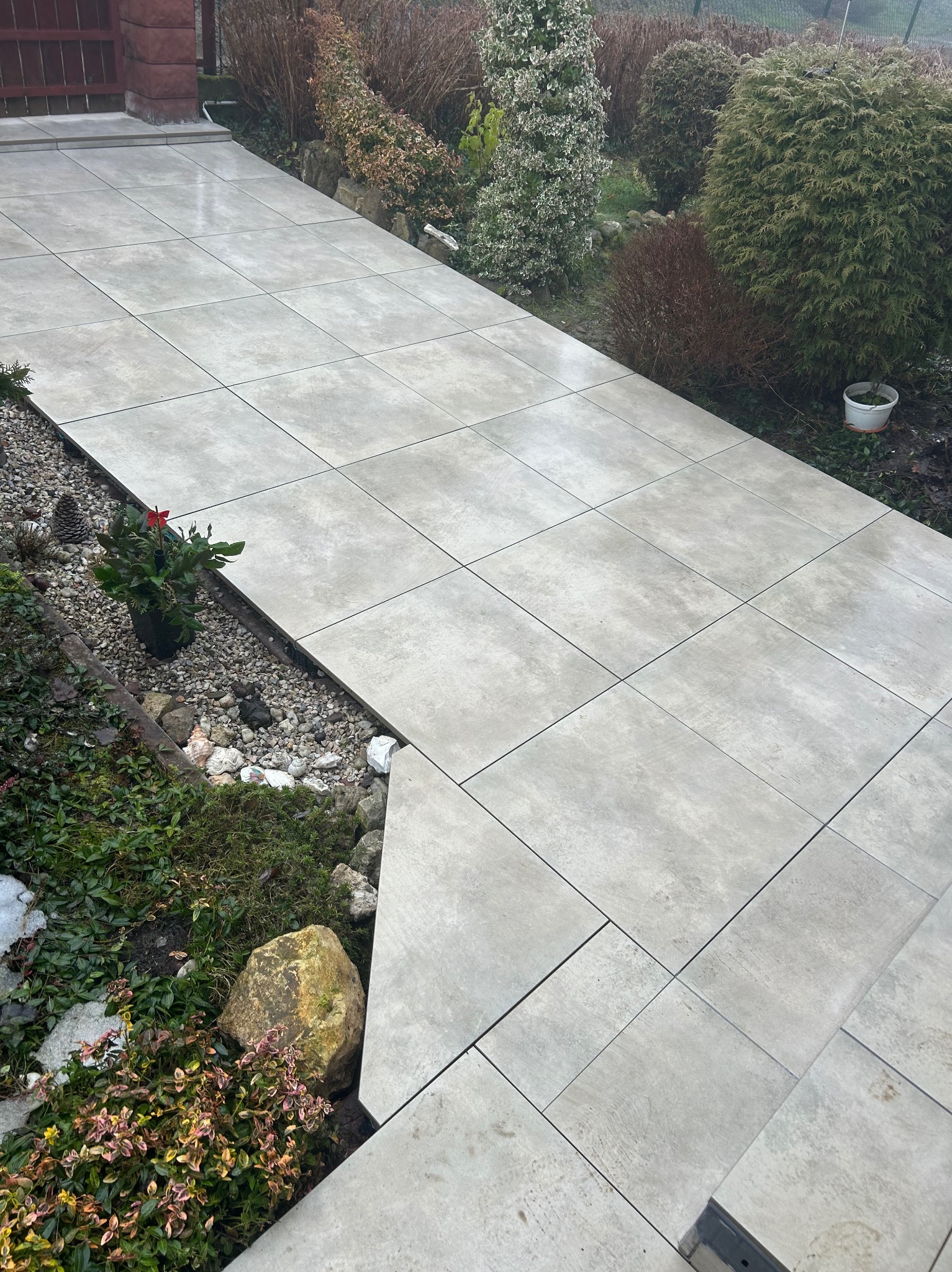Ułożona, jasnoszara kostka brukowa tworząca ścieżkę w ogrodzie, z widocznymi roślinami i kamieniami po bokach. Ścieżka prowadzi do schodów.