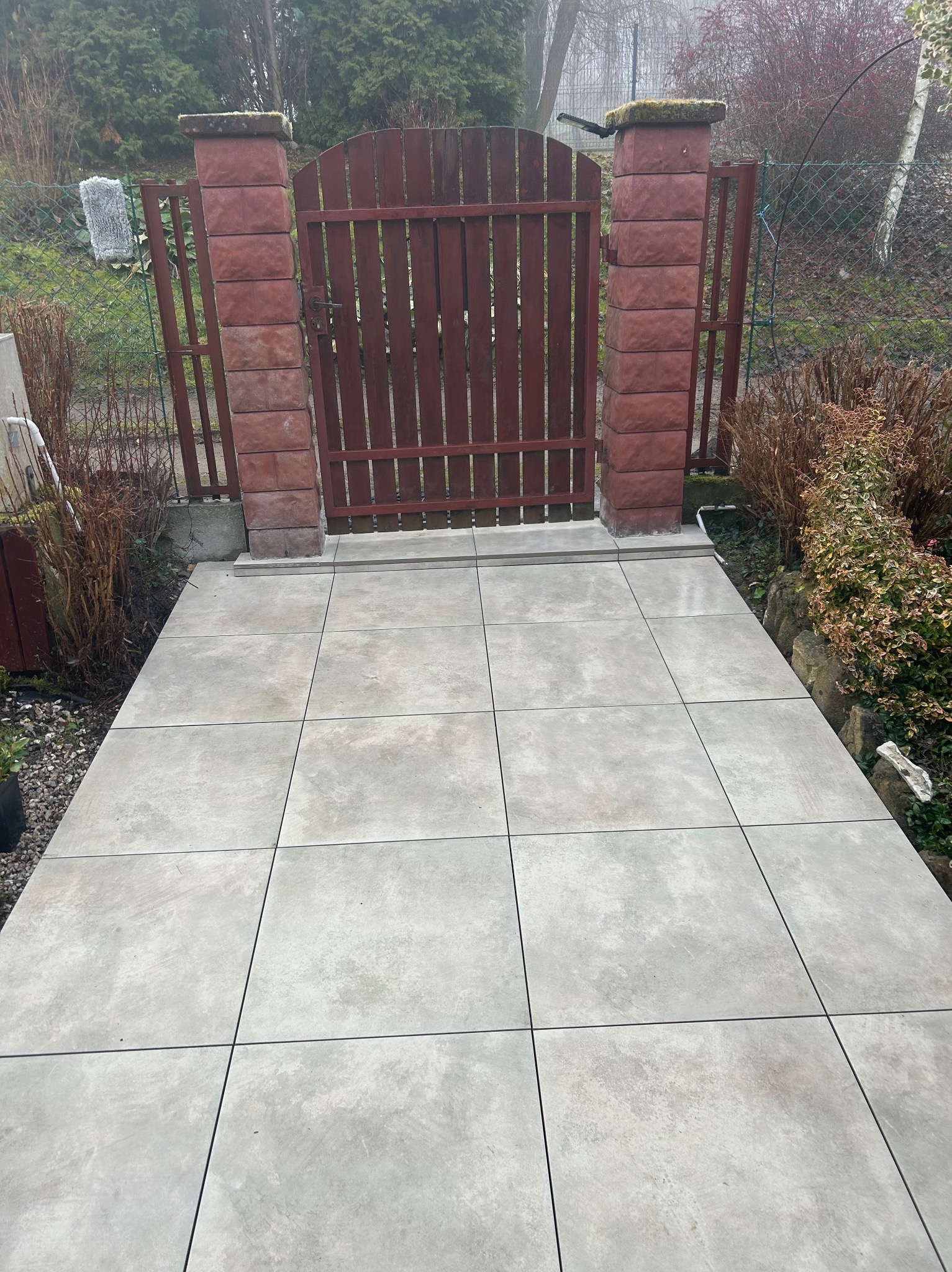 Ułożona, jasnoszara nawierzchnia z płyt prowadząca do drewnianej furtki z ceglanymi słupkami. Widok na ogród w tle, pochmurny dzień.