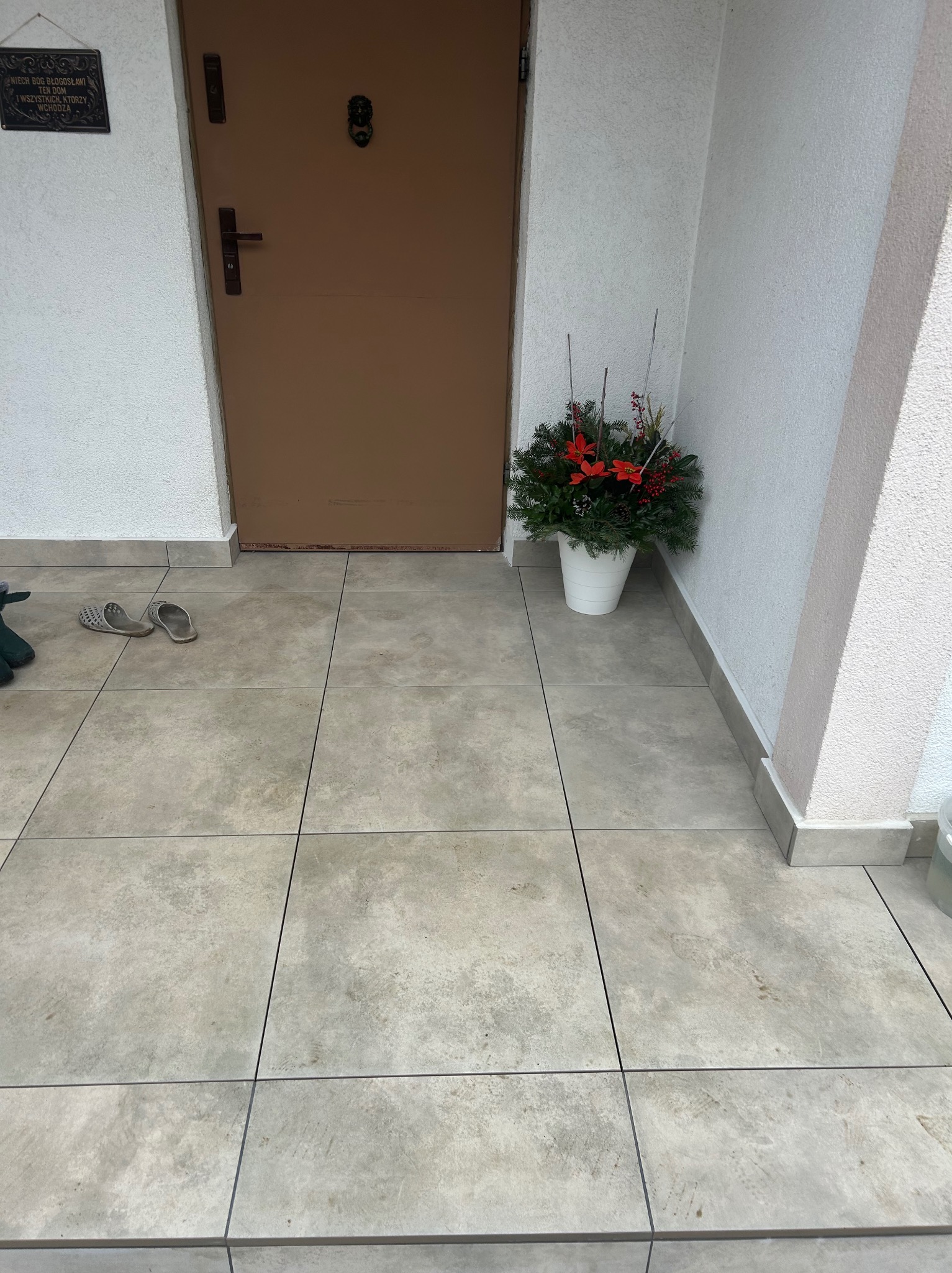 Kafelki na zewnątrz domu, brązowe drzwi wejściowe, dekoracja świąteczna w doniczce, buty robocze i tabliczka z napisem przy wejściu.